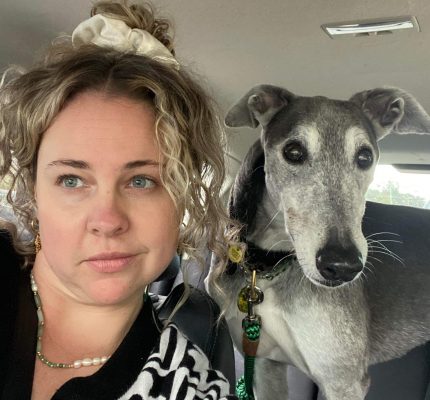 A selfie of a woman with curly hair and a blue greyhound next to her.